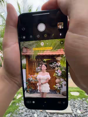 A person's hands holding an iPhone 14 Pro Max, which is displaying a photo of a young girl standing in front of a wooden structure with plants and flowers. The scene is framed as if taking a photo, with the phone's camera UI visible. The background shows a green environment with vegetation. The composition is well-lit and colorful.