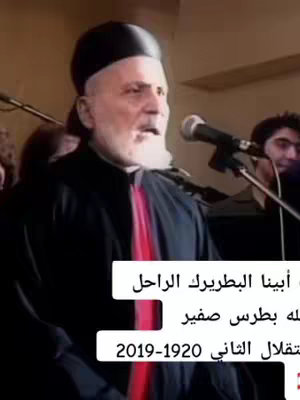 The cover image shows a medium shot of an older man speaking into a microphone. He is wearing a black robe with a red stripe down the front, a black hat, and a white beard. Behind him, other people and a microphone stand are barely visible. Text overlays at the bottom-center provide additional context.