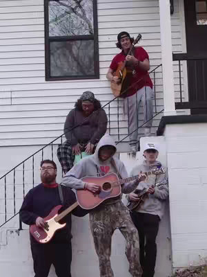 A group of five young men, presumably a band, are posing on the steps of a house. The background is a simple white-sided house with a dark window and a black railing, and the men are holding instruments, including guitars and a bass, as they get ready to play.