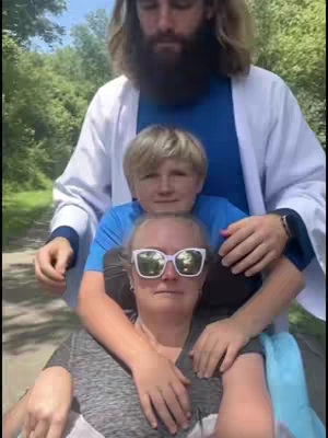 The cover image features three people: a woman in sunglasses and a gray t-shirt, a young boy, and a man appearing to be dressed as Jesus. The man stands behind the boy and the woman, with his hands on their shoulders. The background is a blurry green, suggesting an outdoor setting. The composition has a devotional feel because of the man's attire, suggesting religious themes.
