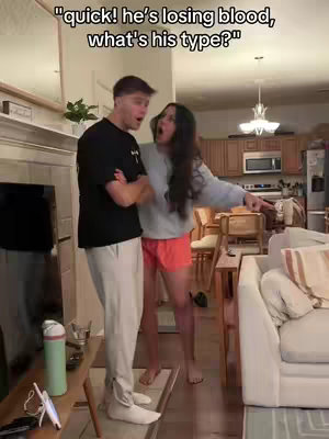 Two people, a man and a woman, are in a home setting. The man is on the left, wearing a black t-shirt and light-colored pants, looking towards the camera with an expression of mild surprise. The woman on the right is wearing a light-colored sweater over a pair of orange shorts, pointing emphatically, also with a surprised expression. The background shows a living room setting with a fireplace, TV, and kitchen area with cabinets and appliances.
