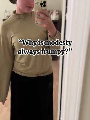 A woman in a light green long-sleeved shirt and a black skirt stands in front of a mirror, holding a phone to take a selfie. Text overlay reads, "Why is modesty always frumpy?"
