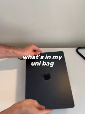 A person's hands holding a closed, black laptop. The laptop is centered against a white background. Text overlayed in white states, "what's in my uni bag".