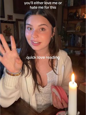 A woman in a white hoodie is holding a deck of cards and gesturing with her left hand. Above her head, text reads "you'll either love me or hate me for this." Below her, text reads "quick love reading." A lit candle stands to the right of the frame.