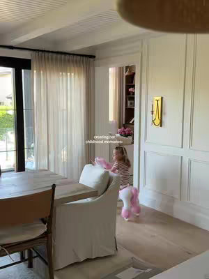 A bright, well-lit interior shot featuring a young girl holding a pink toy pony. The room has large windows with sheer curtains, wood floors, white paneled walls, and a glimpse of a built-in cabinet. The girl is the central focus, positioned in the foreground near a white chair, with the pony clearly visible.