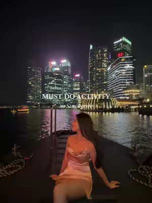 A woman in a silky dress sits on a boat at night, looking away from the camera towards illuminated city buildings. Text overlayed on the top reads, 'MUST DO ACTIVITY in Singapore.' The background includes city lights, a river, and a clear night sky.
