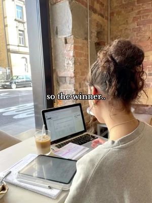 A person is sitting at a table, viewed from behind. They have their hair up in a bun. In front of the person is a laptop with text visible on the screen, a tablet, a notebook, a pen, and a drink with a straw. Beyond the person is a window with visible street scene.