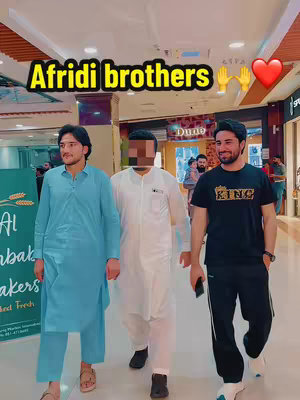 Three men walking in a mall setting. One is wearing a blue suit, one a white outfit and the third, wearing a black shirt that says 'KING', and black pants. The background shows a shopping mall with other people.