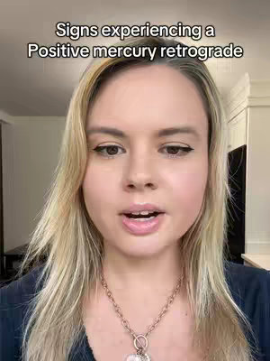 A woman with long blonde hair and a dark blue shirt looks directly at the camera, speaking. Text overlays the image saying, 'Signs experiencing a Positive mercury retrograde.' The background is a blurred indoor setting with a white wall.