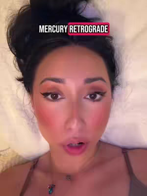 A close-up shot features a woman with dark hair pulled up, wearing makeup, and lying on a white surface. The image includes text overlayed at the top that states "MERCURY RETROGRADE". with her mouth slightly open, indicating she is likely speaking. The lighting is bright, and the focus is primarily on her face.