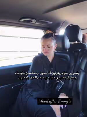 The image shows a person sitting inside a car, likely in a back seat. The person, who appears to be a woman with light skin and hair tied up in a bun, is wearing a black hooded top and has a somewhat contemplative expression. She seems to be looking down and to the side. Above her, the car's interior is visible, including a window and part of the ceiling. Text overlays are added above her head and at the bottom of the image, providing context or commentary. The overall composition suggests a candid moment or reaction.