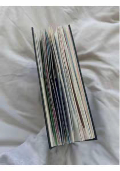 An open book is lying on a white, crumpled surface. The book is dark-colored with pages that are visible and slightly fanned out. The pages have various colors and are oriented to the left.