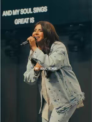 A woman singing into a microphone on stage. She is wearing a light denim jacket and jeans against a dark background.