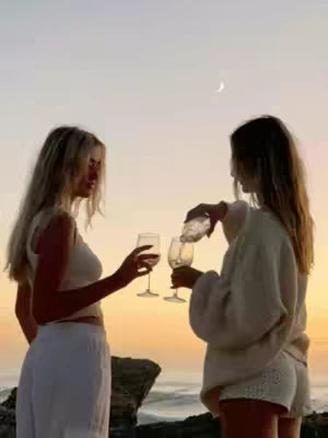 Two women are at the beach during sunset. One is pouring what looks like white wine into a glass held by another woman. Both women are fair-skinned with long blonde hair. The sky is yellow and pink.
