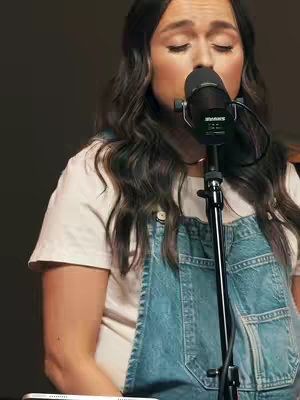 A woman with long, dark hair is singing into a microphone. She wears a white t-shirt and denim overalls. Her eyes are closed, and her expression is focused. The background is dark and out of focus. A microphone stand is positioned in front of her.