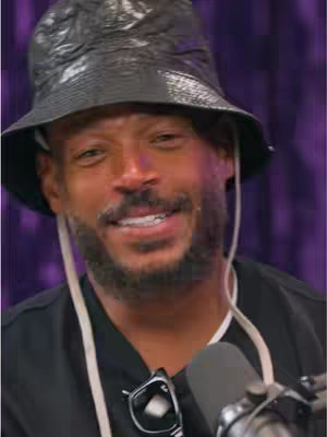 The cover image features a close-up of Marlon Wayans wearing a black bucket hat and a black shirt. He has a slight smile and a neatly trimmed beard. A microphone is visible in the foreground, and a pair of glasses hangs from his shirt. The background is a blurred purple color.