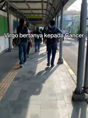 The image captures a public transit scene with several people walking on a tiled pathway under a covered walkway. The walkway is flanked by a wall on one side and what appears to be a road on the other. The lighting suggests it might be daytime, with sunlight casting long shadows from the support pillars. The text overlayed on the image reads, 'Virgo bertanya kepada Cancer'.