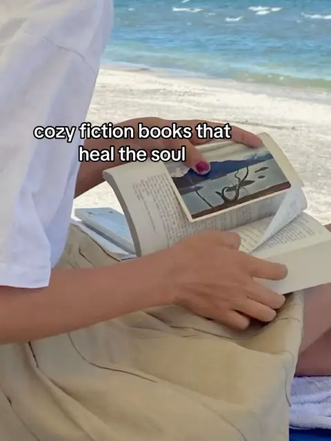 A person is sitting on a sandy beach, reading a book and enjoying the view of the clear blue sea on a summer day. The person's hands and torso are visible; they are wearing a white t-shirt and a beige skirt. Text overlays the image.