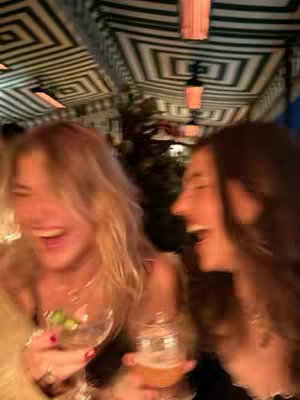 Two women are laughing at a restaurant, holding drinks. The background is blurred, but appears to be a tent-like setting with a repeating geometric pattern on the ceiling.