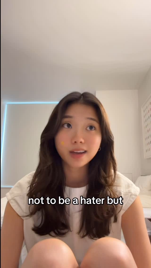 A young woman with long brown hair is centered in a selfie-style video. She's wearing a white top and has a small yellow sticker on her cheek. The background includes a plain wall, a glimpse of a bed, and a soft light fixture.