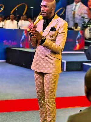 A man in a patterned suit stands on a stage, holding a microphone. The background has colorful lighting and some individuals