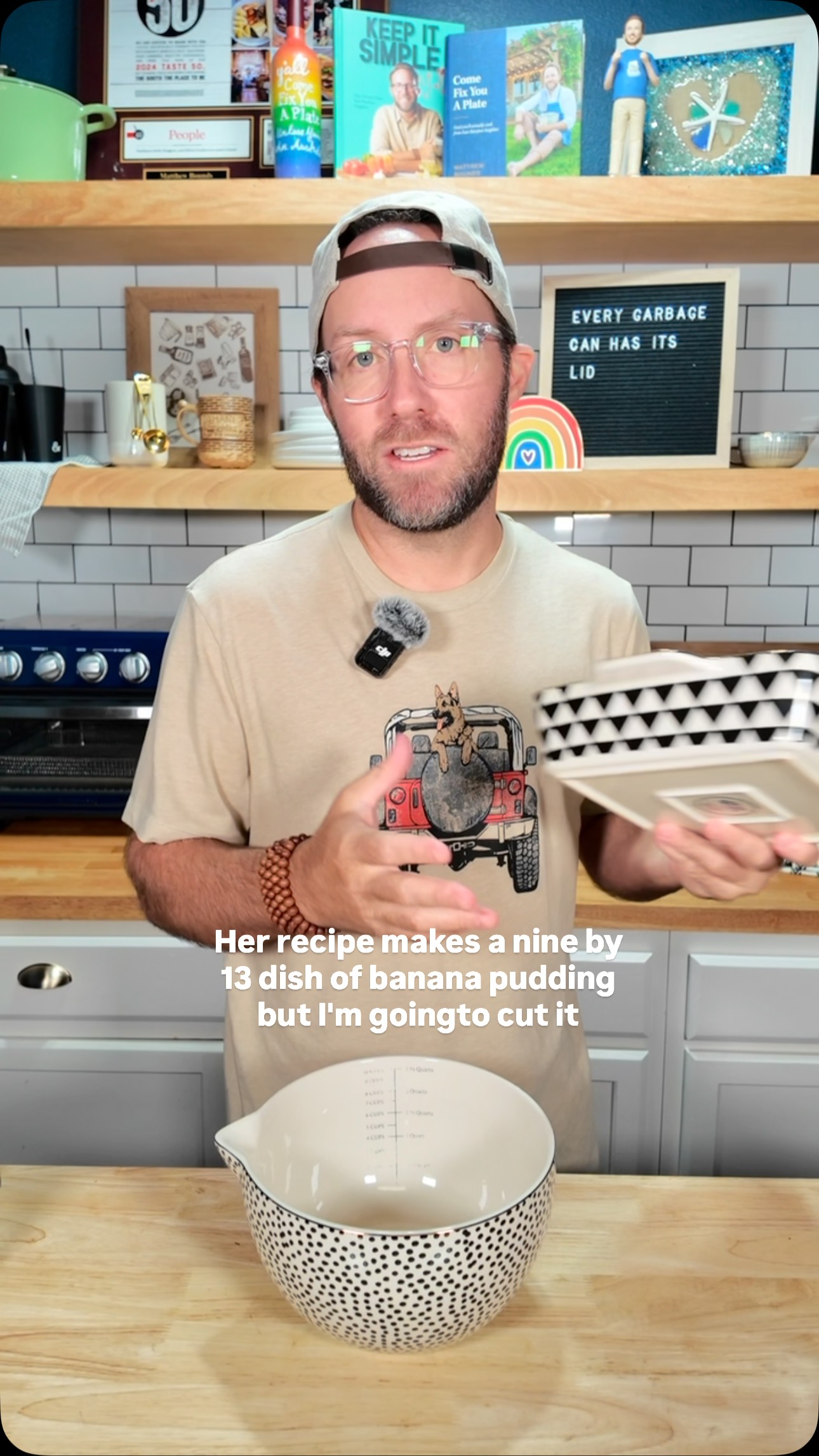A man with a beard and glasses stands in a kitchen, holding a square baking dish. He is wearing a light brown t-shirt, a baseball cap, and a microphone is attached to his shirt. The background has a white tile backsplash and wooden shelves stocked with various kitchen-related items. A large black and white polka-dotted mixing bowl sits on a wooden countertop in front of him.