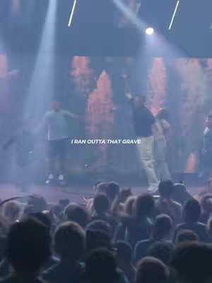 The image shows a stage with spotlights shining down. Several figures are on stage with their hands raised. A crowd is visible in the foreground.