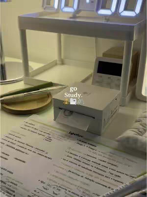 A still-life shot of a desk setup, focused on a printer next to some study notes. The scene is bathed in soft, warm lighting. There's a white desk with a shelf above it, on which digital clocks are placed. A pen and small wooden tray are on the desk in front of the printer, and in the background is an additional studying material. The entire scene has a clean, organized aesthetic.