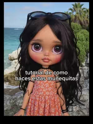 The image is centered on a doll with large, expressive eyes and black hair, accessorized with sunglasses atop its head. The doll wears an orange floral dress, set against a blurred background of what appears to be a beach scene. The composition focuses on the doll, and the text overlay at the bottom in white indicates instructions on making the doll.