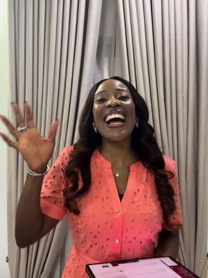 A woman with dark hair and a coral-colored dress is smiling broadly, waving with one hand. She appears to be inside, with beige curtains in the background. She is holding a partially visible tablet or notepad.