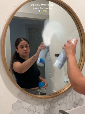 A woman in a black top is spraying cleaning solution onto a large round mirror; her reflection and the spray are also visible. The mirror hangs above a white and gray tiled backsplash.