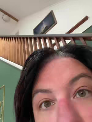 Close-up shot of a person's face, focusing on their eyes and forehead area. They are positioned in a domestic setting, with a staircase, artwork, and a light visible in the background. The background is out of focus.