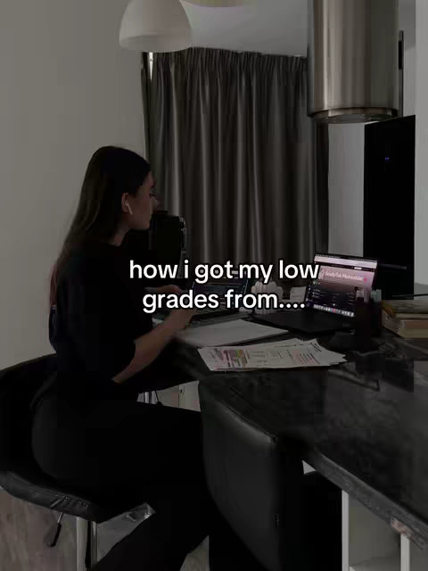 A woman is sitting in front of a laptop, leaning against a kitchen counter. Text on the screen appears to be some type of note-taking app, and there is paper on the counter as well. The lighting is soft, and the room is tidy. The background of the image consists of a dark-colored curtain, a kitchen cabinet, and a ceiling light. The woman is wearint an all-black outfit.