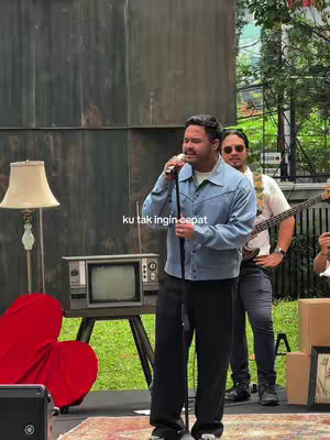 A man is singing into a microphone, with a band playing in the background. The setting appears to be outdoors, with a vintage television, a retro, heart-shaped chair, and greenery.