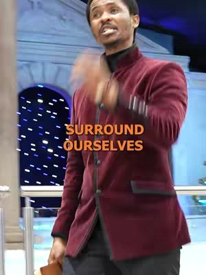 A man in a burgundy jacket with a black turtleneck speaks, raising his hand in a gesture towards the right; text in the image reads, 'SURROUND OURSELVES'. The background has architectural features and blue and white lighting details.