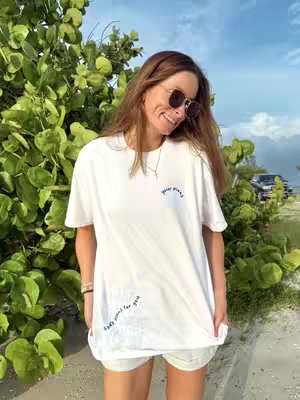 A woman with long brown hair wearing sunglasses and a white t-shirt, stands outside in front of some green bush. The woman is smiling while looking to the left of the camera. In the background there's a clear blue sky, indicating the sun. The t-shirt contains the text "your plans" and "God's plans for you".