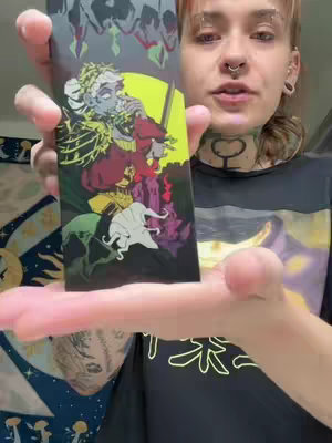 A person with multiple piercings and tattoos holds up a tarot card displaying an illustration of figures in a colorful setting and blurred background.