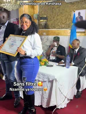A woman in a white blouse and blue jeans holds a framed certificate, smiling at the camera. She is standing. Several people are around a table with a white tablecloth, and a Congolese flag can be seen in the background.