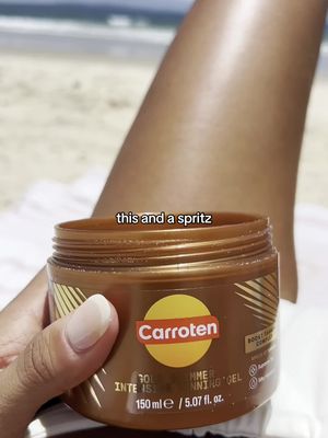 The image shows a person's tanned leg resting on a beach towel, with an open jar of Carroten tanning gel in the foreground. The surrounding area has sand and ocean visible in the background. The composition is focused, with the tan leg taking up most of the frame, and golden brown accents of the jar.