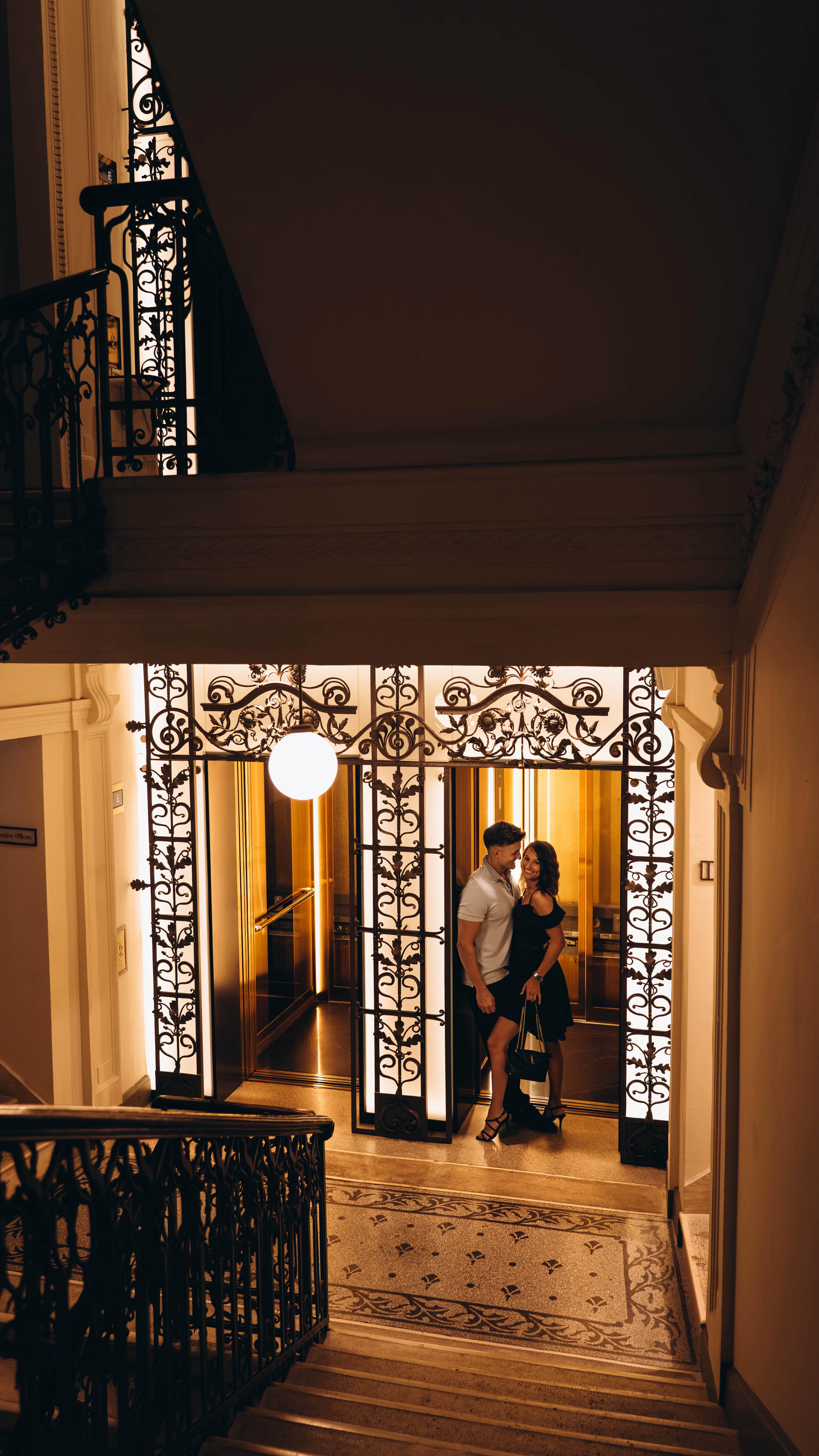 A couple embraces at the entrance to a hotel, a grand doorway with ornate ironwork and ambient lighting creating a warm glow. The composition is from a low angle looking up, highlighting the architectural detail and the romantic moment.