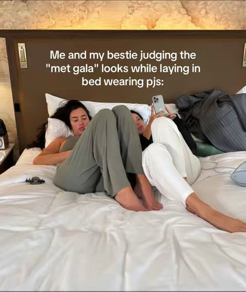 Two women are lying in bed, one is holding a phone and taking a photo, the other is looking up at the phone. The image is brightly lit and seems casual. The background has a dark brown headboard with gold details adding a touch of luxury. The sheets are white, making the scene clean and comfortable. The text on the image gives context: "Me and my bestie judging the "met gala" looks while laying in bed wearing pjs:".
