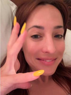 A close-up shot of a woman's face, angled slightly upwards. She has fair skin, auburn hair, and is lying on a white sheet or pillow. Her right hand is raised towards the camera, with her fingers spread, displaying bright yellow, long, pointed fingernails. She is smiling gently, with a subtle expression. The lighting is soft and diffused.