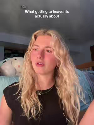 A young woman with long, wavy blonde hair is the focus. The text "What getting to heaven is actually about" is at the top, written in a simple, sans-serif font. The woman looks slightly up and to the left, with a somewhat sad or contemplative expression. The background is indistinct, showing a hint of a room with a bed and pillows, and the lighting appears to be soft and natural.