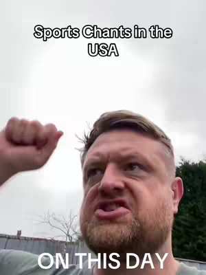A man with short, light-brown hair makes a facial expression of surprise and frustration, his mouth slightly open, one hand in a fist. Text at the top says "Sports Chants in the USA". The background is a blurry, overcast sky and some green foliage.