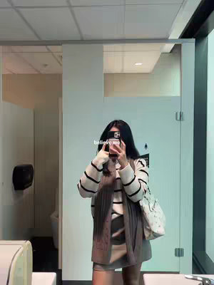 A woman is taking a mirror selfie in what appears to be a public washroom. She is holding a phone up to her face to capture the image, with her face obscured. She is dressed in a striped sweater, with a scarf and a purse over her shoulder. The setting includes mirrors, restroom stalls, and overhead lighting.