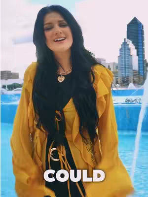 A woman with long black hair, wearing a yellow top, stands in front of a fountain with blurred background of city buildings. The hook says 'COULD'.