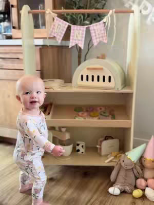 A young child stands in front of a wooden play kitchen, looking directly at the camera. She is wearing patterned pajamas. The kitchen has multiple shelves filled with toy accessories. Beside the child are stuffed animals wearing party hats . A banner that reads “ONE” hangs above the play set.
