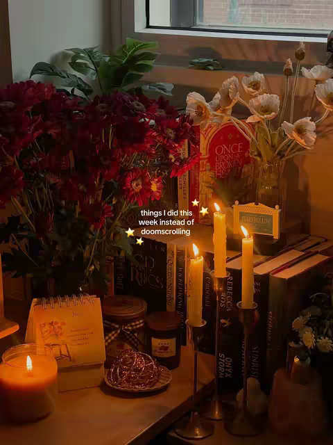 A warm, inviting indoor scene featuring various objects arranged on a surface, likely a table or shelf. The composition is dominated by a vibrant bouquet of dark red flowers to the left, paired with white flowers in a vase. Books are stacked in the background, suggesting a cozy atmosphere. Several lit candles of varying heights add warmth and ambiance. Additional items include a calendar, small jars, and decorative elements, all contributing to a visually rich and layered setup. The lighting is soft, creating a sense of intimacy.