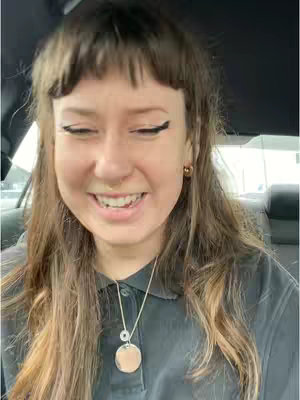 A selfie-style shot featuring a young woman smiling broadly. She has light brown hair with bangs and is wearing a dark top and a necklace. The background is blurred, suggesting she is in a car.