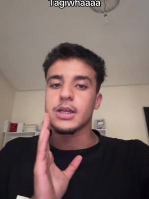 The image features a young man in a black shirt looking directly at the camera, with his right hand raised, fingers spread out near his face. The background is a plain, light-colored wall, with what appears to be a section of a shelf holding some items in the upper left corner, and a ceiling light in view. A text overlay is visible above his head.